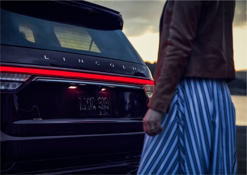 A person is shown near the rear of a 2023 Lincoln Aviator® SUV as the Lincoln Embrace illuminates the rear lights | LaFontaine Lincoln Flushing in Flushing MI