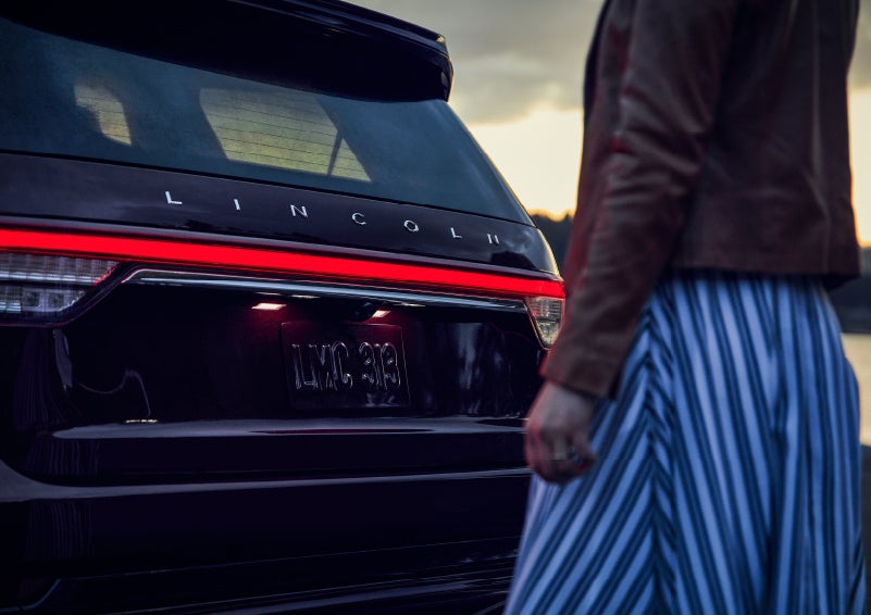 A person is shown near the rear of a 2024 Lincoln Aviator® SUV as the Lincoln Embrace illuminates the rear lights | LaFontaine Lincoln Grand Blanc in Flint MI