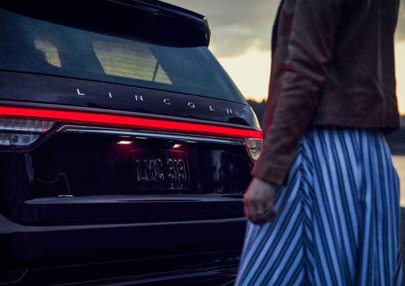 A person is shown near the rear of a 2024 Lincoln Aviator® SUV as the Lincoln Embrace illuminates the rear lights | LaFontaine Lincoln Flushing in Flushing MI