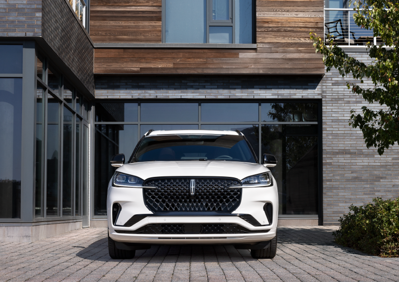 A white 2025 Lincoln Aviator® SUV is shown parked outside a modern building with gray brick and wood accents. | LaFontaine Lincoln Grand Blanc in Flint MI