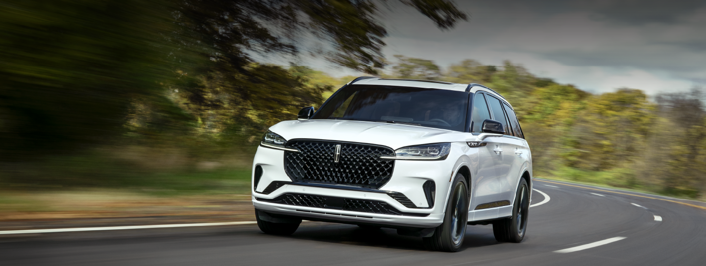 A white 2025 Lincoln Aviator® SUV is being driven down a road lined with trees. | LaFontaine Lincoln Grand Blanc in Flint MI