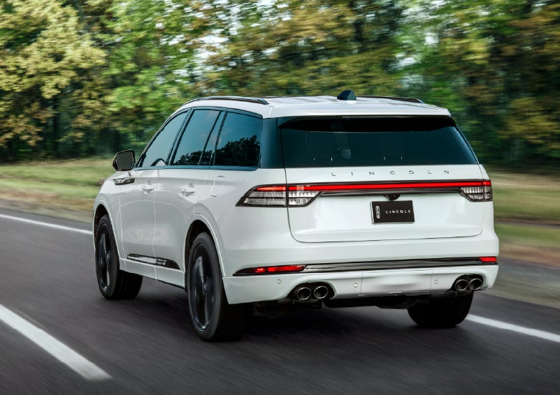A white 2026 Lincoln Aviator® SUV is being driven away down a road lined with colorful trees. | LaFontaine Lincoln Grand Blanc in Flint MI